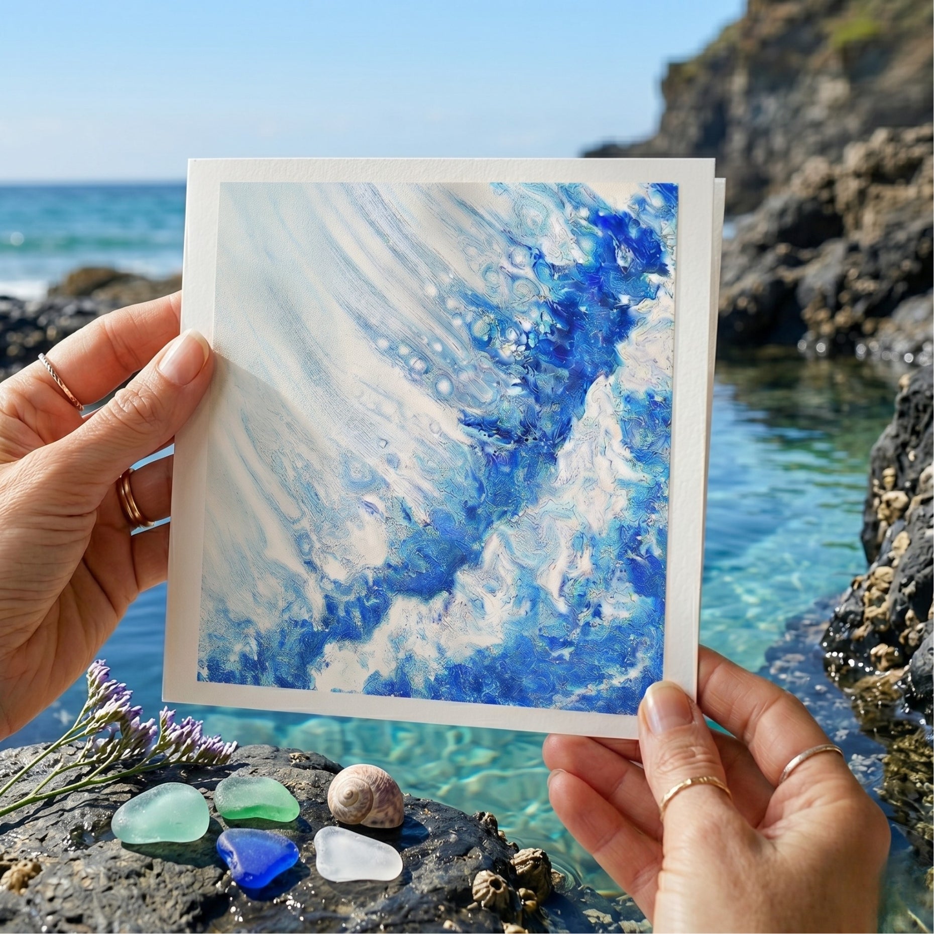 Person holding a small painting of blue and white abstract design by the sea.