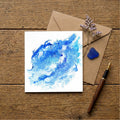 Blue abstract painting on a white card with a brown envelope, pen, and dried flowers on a wooden surface.