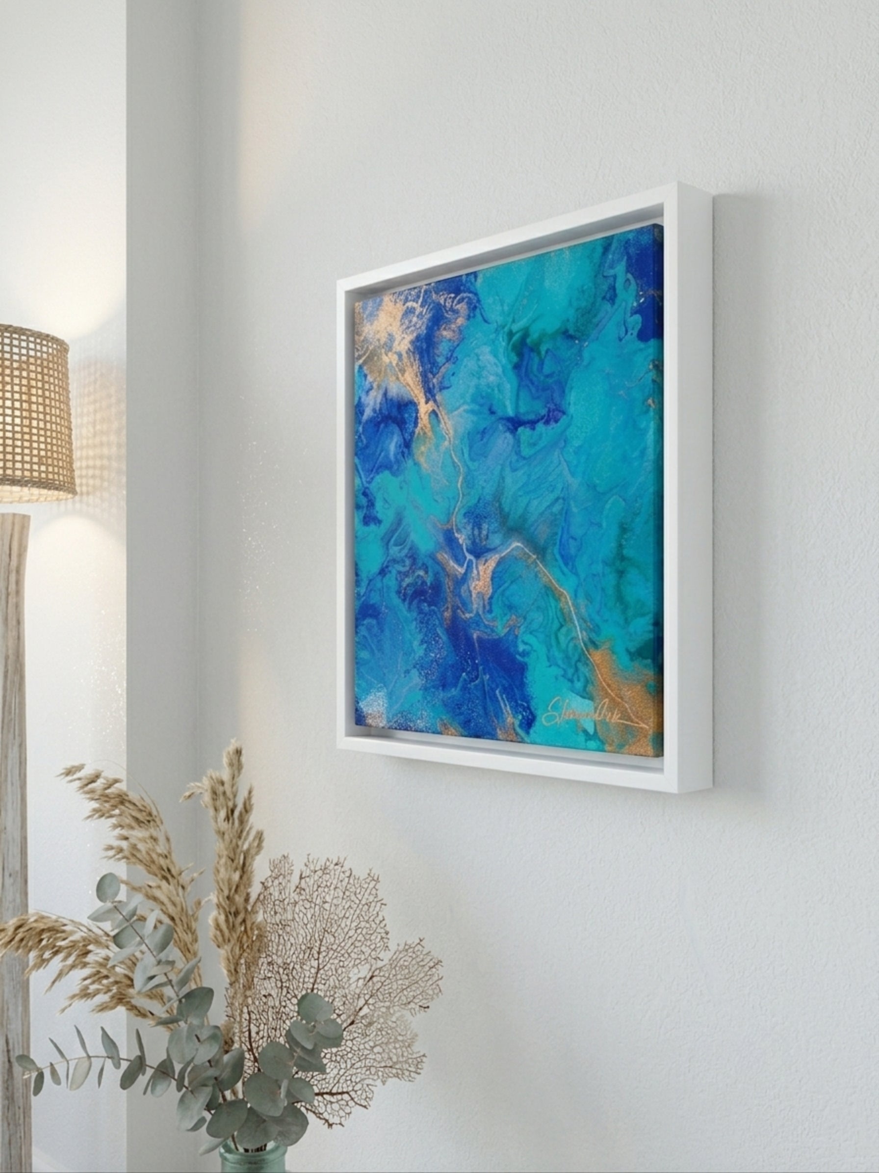 Framed abstract artwork with blue and gold tones on a white wall, next to a lamp and plant.