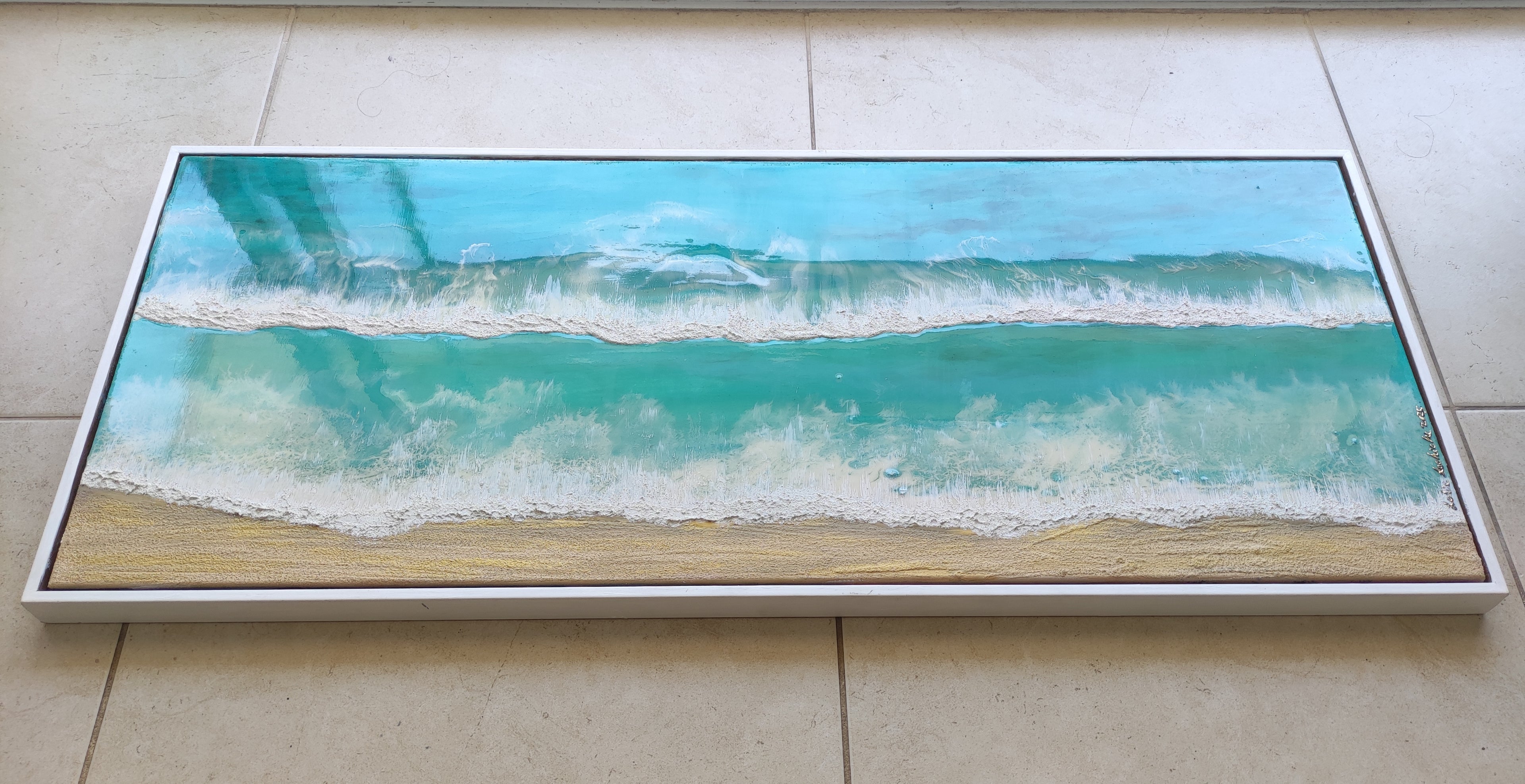 Framed artwork of a beach scene with blue water and white waves on a tiled floor.